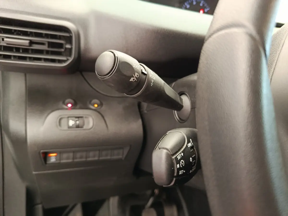 Gros plan sur la commande des feux et régulateur de vitesse à gauche du volant dans l’habitacle noir du Peugeot Partner blanc.
