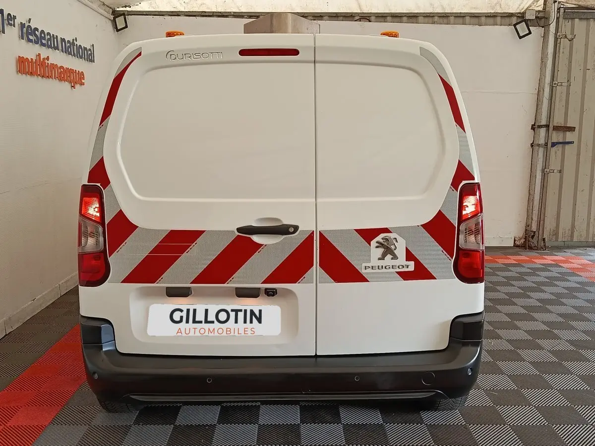 Vue arrière d’un Peugeot Partner blanc avec bandes réfléchissantes rouges et blanches et logo visible sur porte arrière.