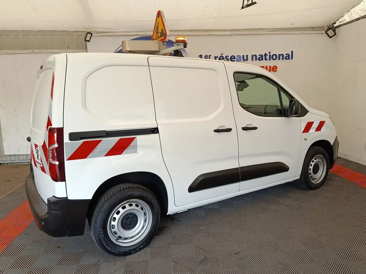 Vue 3/4 arrière droite d’un Peugeot Partner blanc 2022 avec bandes réfléchissantes rouges et gyrophare orange.