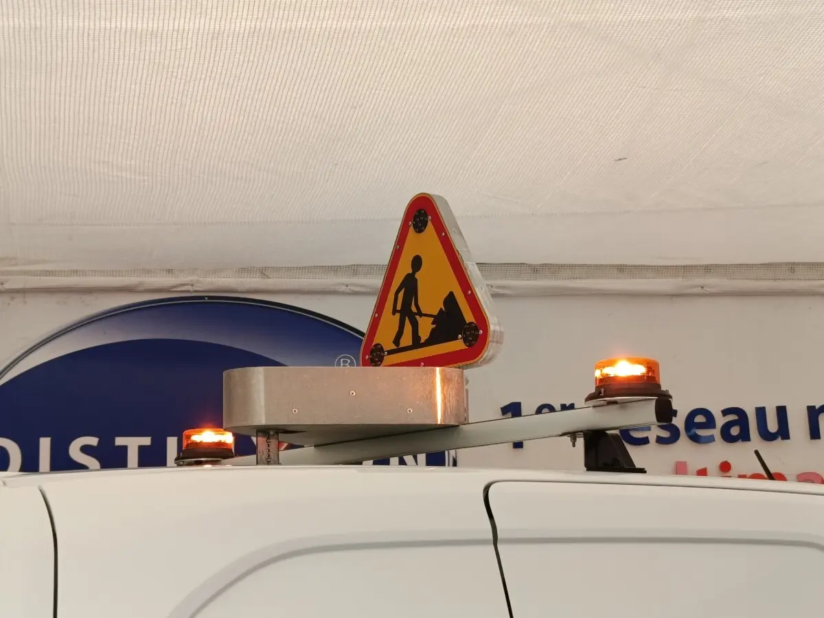 Vue partielle du toit blanc du Peugeot Partner 2022 avec panneau triangulaire de chantier et balises lumineuses orange allumées.