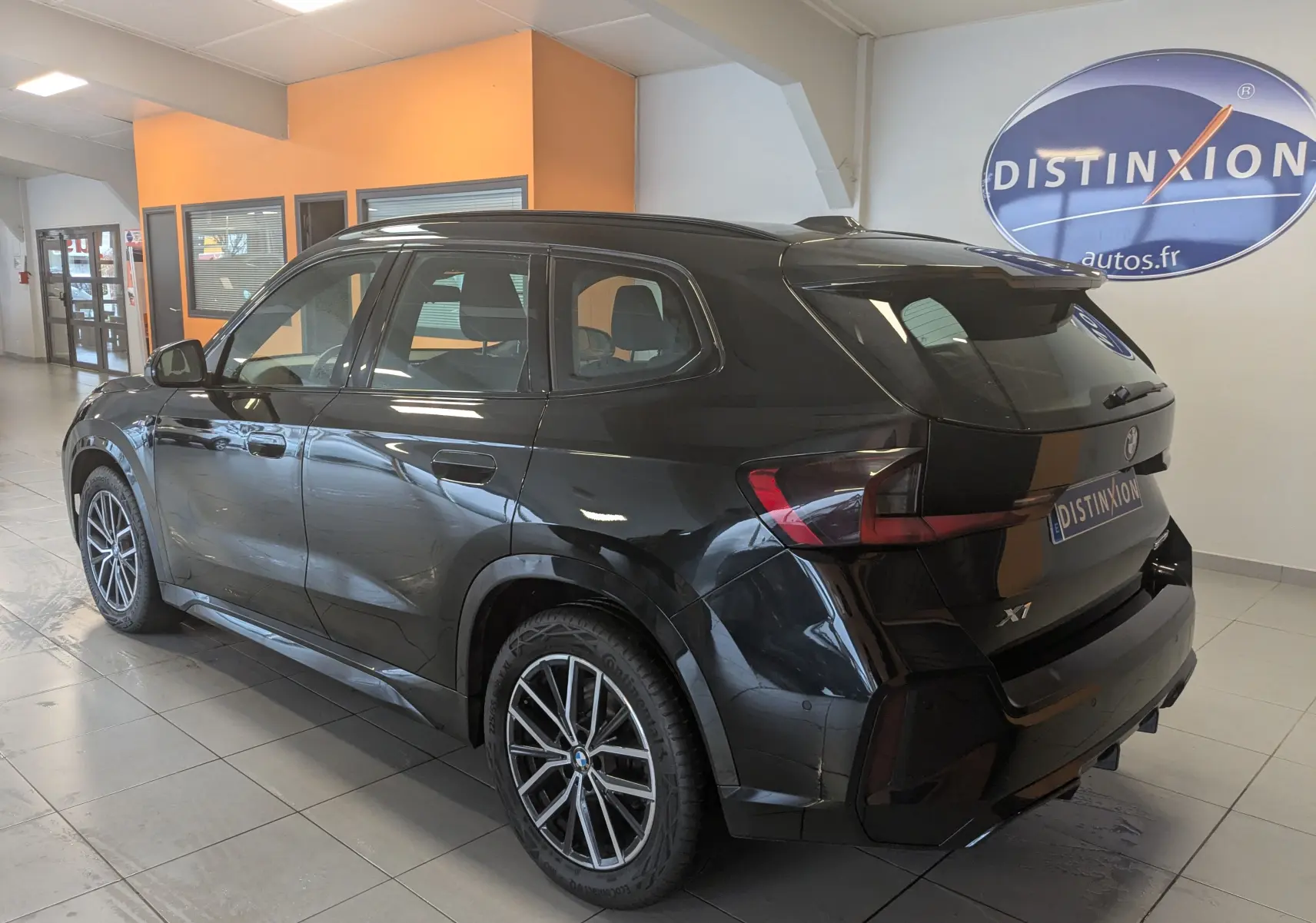 BMW X1 noir vue 3/4 arrière droit en intérieur, avec jantes bicolores et toit ouvrant panoramique.