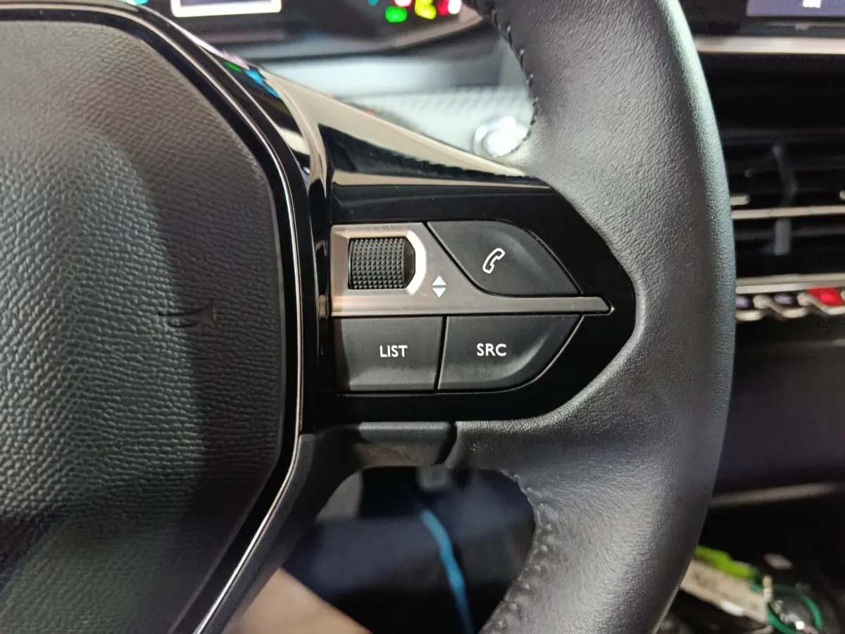 Gros plan sur les commandes gauche du volant cuir d'une Peugeot 208 électrique blanche, avec boutons multifonctions et molette.