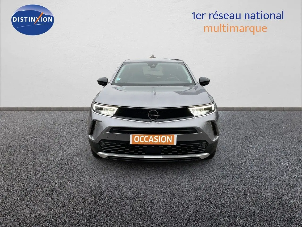 Vue frontale d'un Opel Mokka gris Artens métal avec phares allumés et plaque "OCCASION" visible.