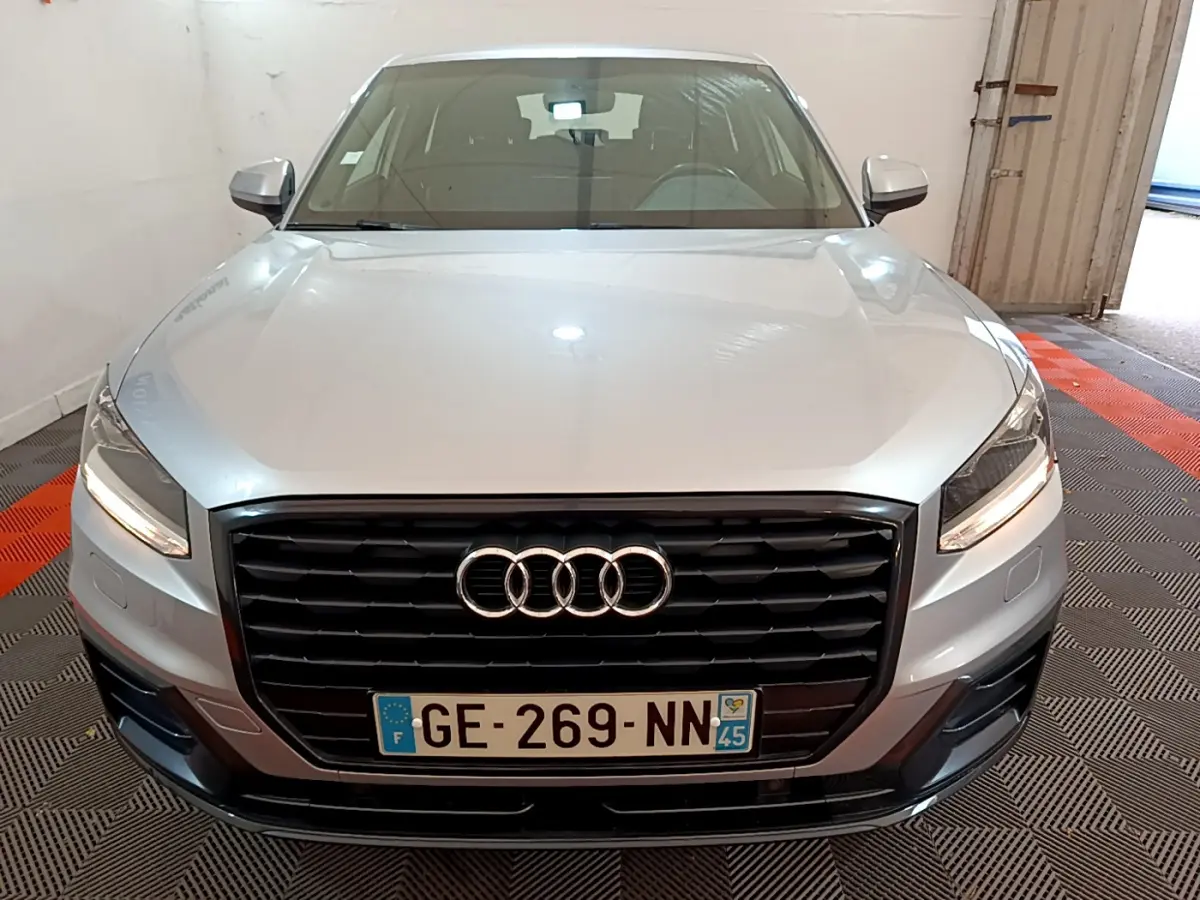 Vue avant d’un Audi Q2 gris clair 2020 avec calandre noire et phares LED allumés dans un garage.