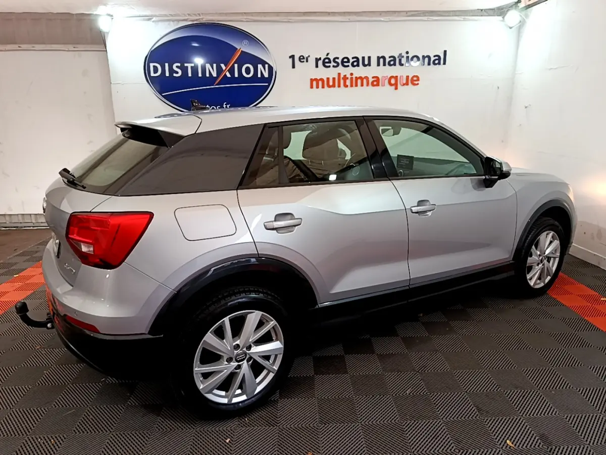 Audi Q2 gris clair vue de côté droit arrière, avec toit noir et feux arrière rouges en intérieur showroom.