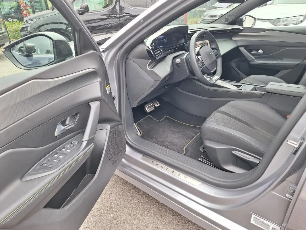 Vue intérieure côté conducteur d’une Peugeot 308 Hybrid 145 e-DCS6 GT gris, avec tableau de bord numérique et sellerie noire.