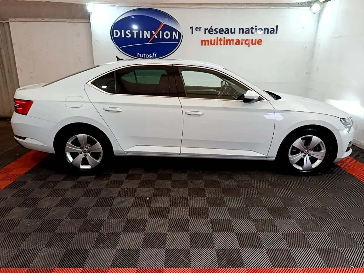Vue de profil côté gauche d'une Skoda Superb blanche 2021 dans un showroom avec sol à damier noir et rouge.