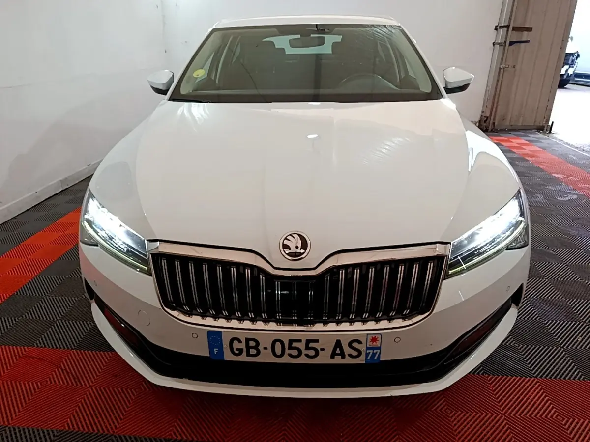 Vue avant d'une Skoda Superb blanche 2021 avec phares LED allumés et calandre chromée dans un garage.