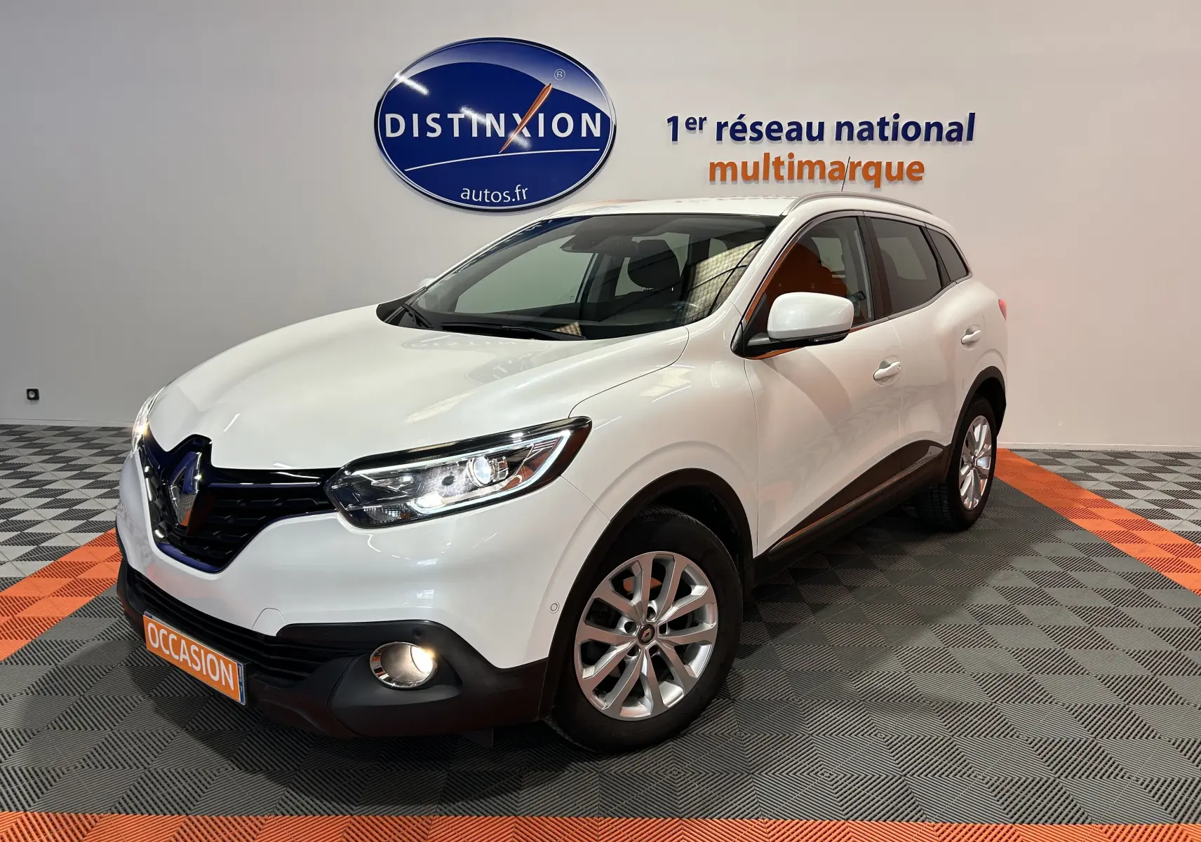 Vue 3/4 avant droit d'un Renault Kadjar blanc avec feux allumés dans un showroom multimarque.
