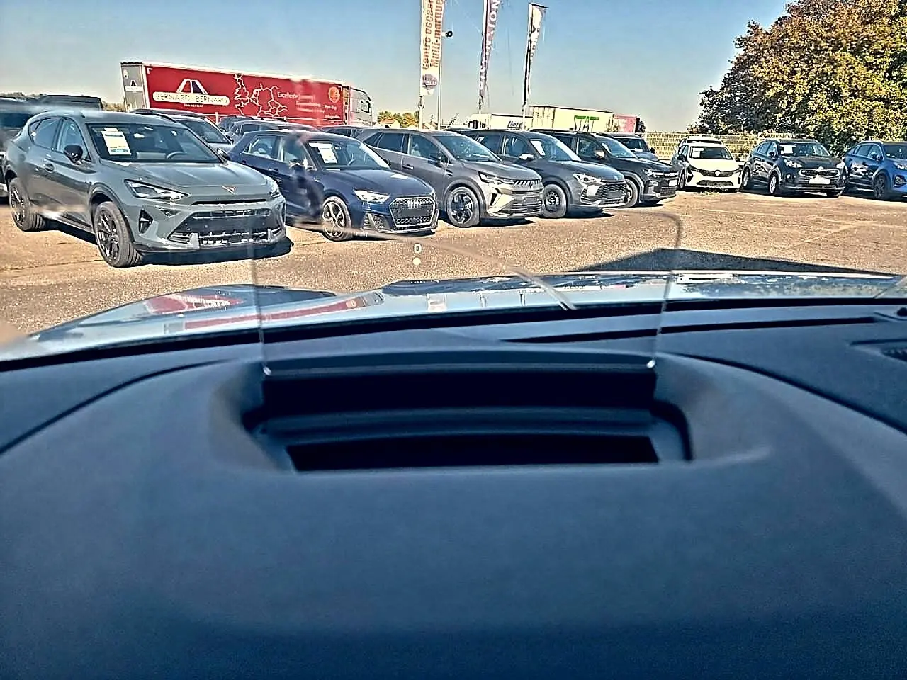Vue depuis l'habitacle vers l'avant, tableau de bord et pare-brise, avec plusieurs voitures en arrière-plan sur un parking.