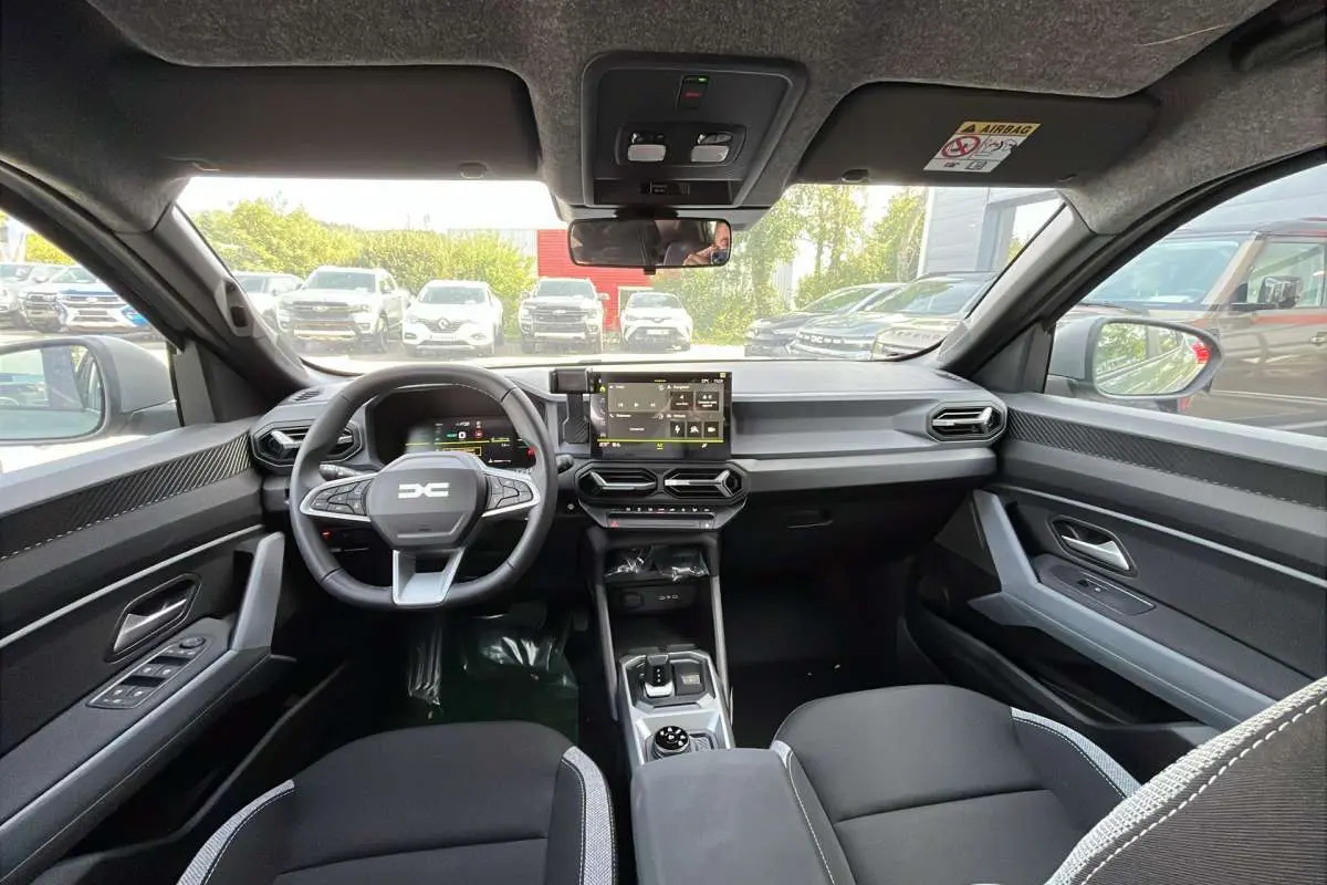 Vue intérieure avant du Dacia Duster Hybrid-G 150 4x4 Journey City, tableau de bord moderne et sièges noirs.