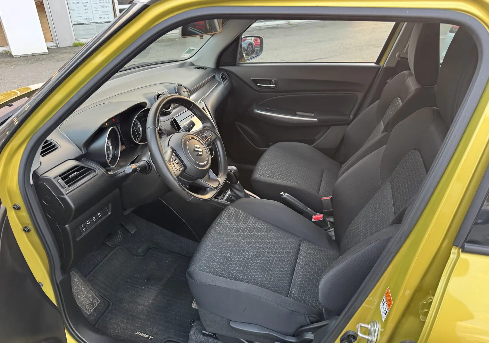 Intérieur côté conducteur d'une Suzuki Swift 1.2 Pack Auto 2023 jaune/gris, avec sièges noirs et tableau de bord moderne.