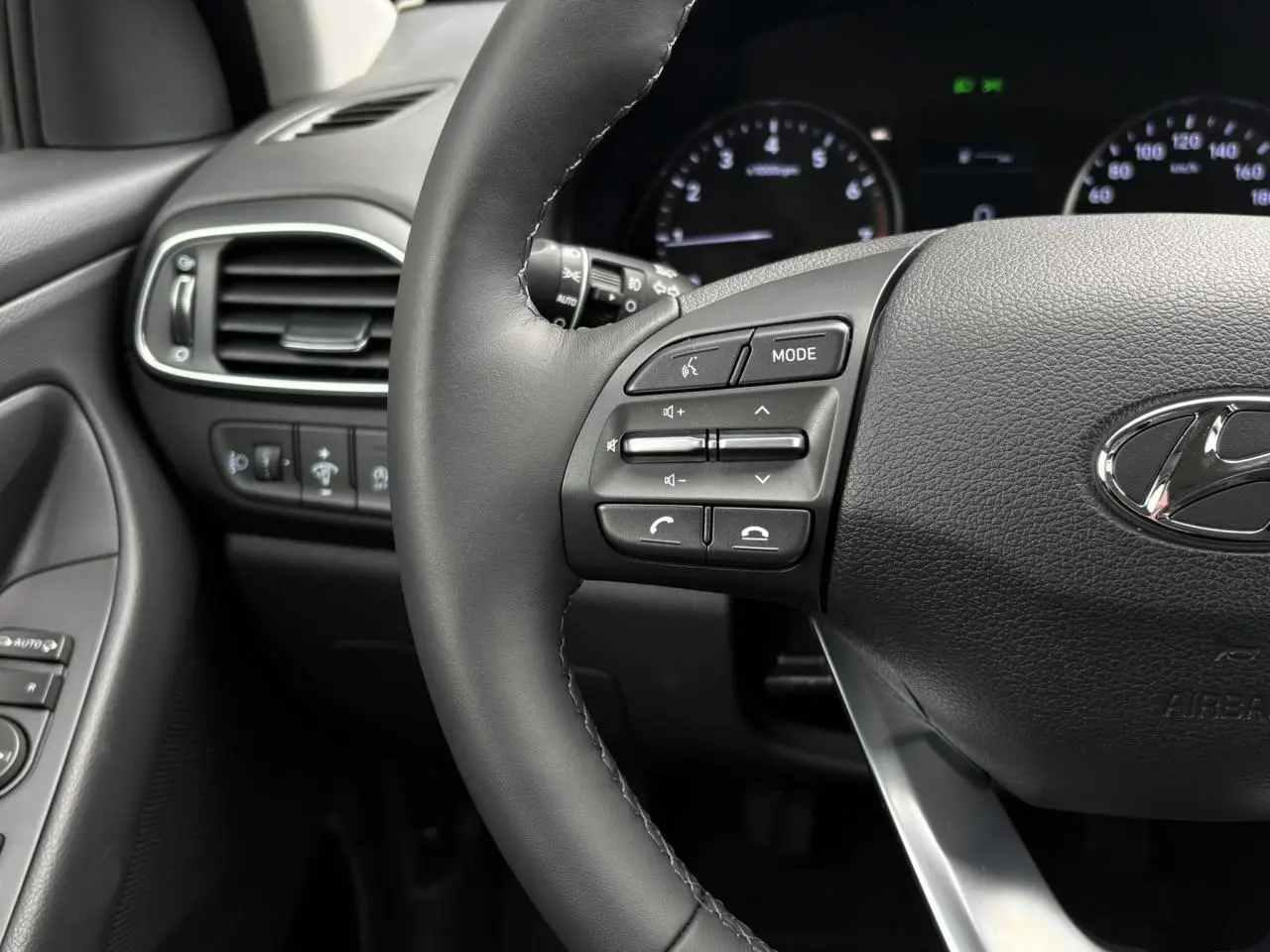 Gros plan sur le volant cuir gris foncé de la Hyundai i30 2025, avec commandes audio et tableau de bord en arrière-plan.