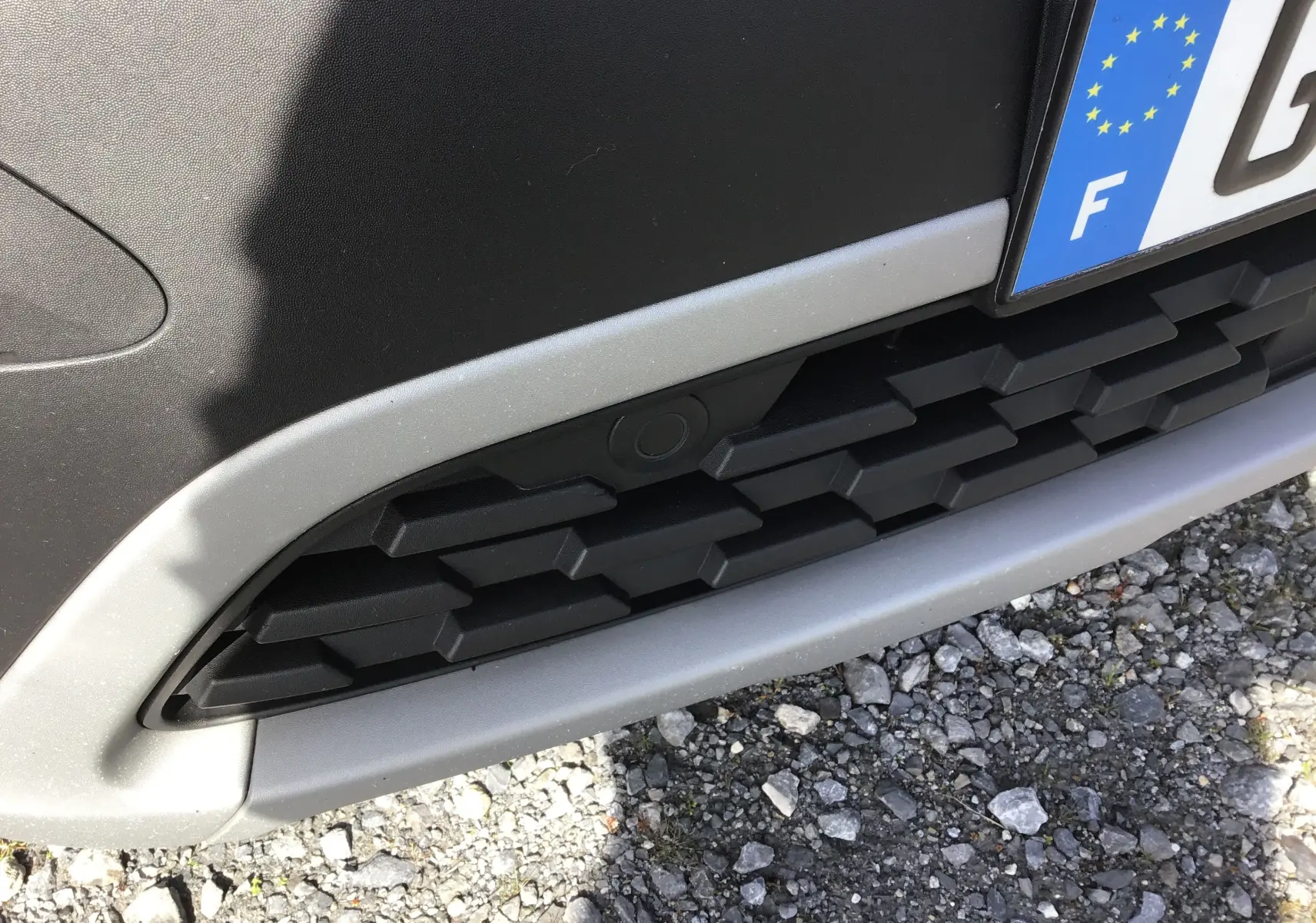 Gros plan sur la calandre avant noire et grise du Renault Kangoo blanc, avec capteur de stationnement intégré près de la plaque d'immatriculation.