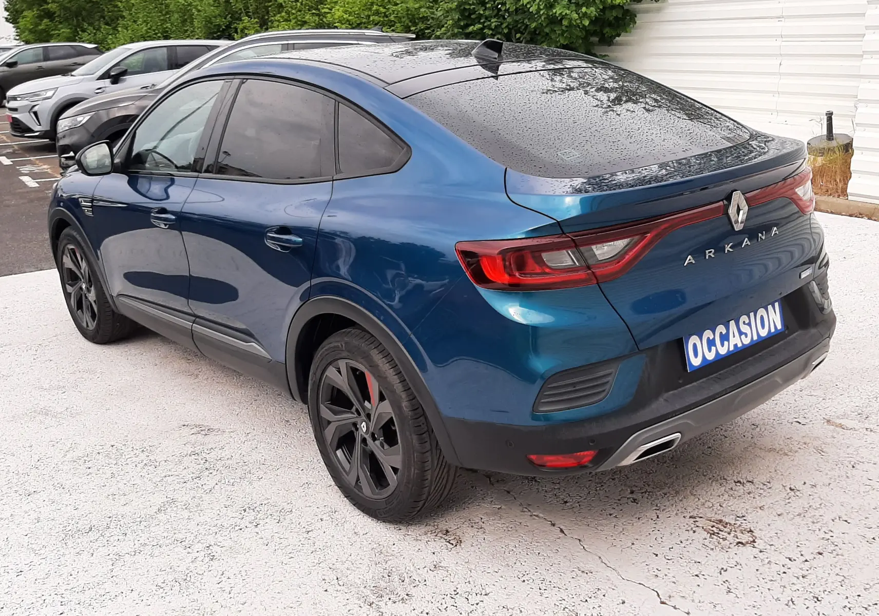 Vue 3/4 arrière droite d'un Renault Arkana E-Tech bleu Zanzibar avec toit noir et jantes noires, stationné sur sol clair.