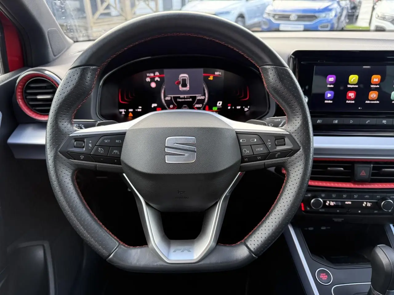 Vue rapprochée du volant sport en cuir perforé noir avec surpiqûres rouges et tableau de bord digital d'une SEAT Arona rouge 2024.