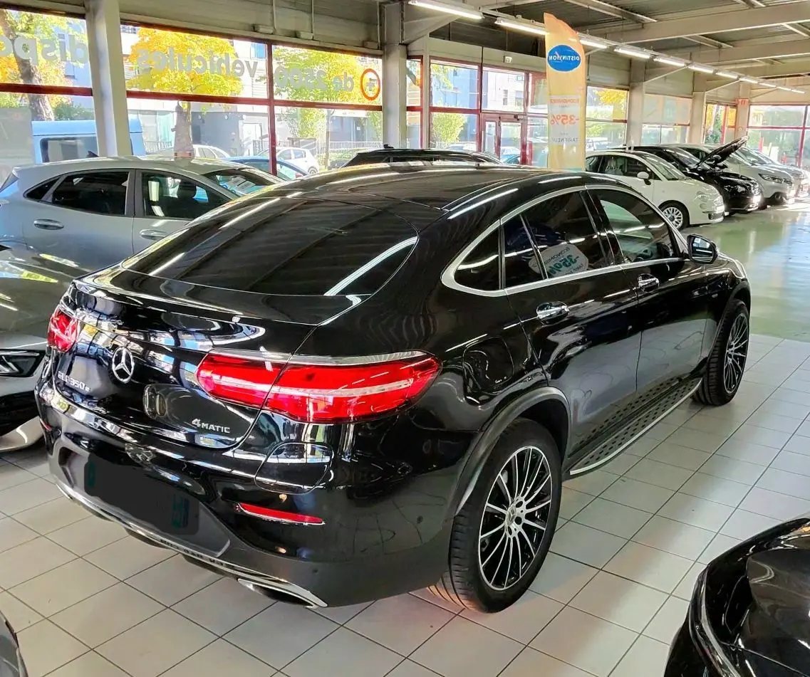 Vue 3/4 arrière droite d’un Mercedes GLC Coupé 350 noir brillant avec jantes sport et feux arrière allumés en showroom.