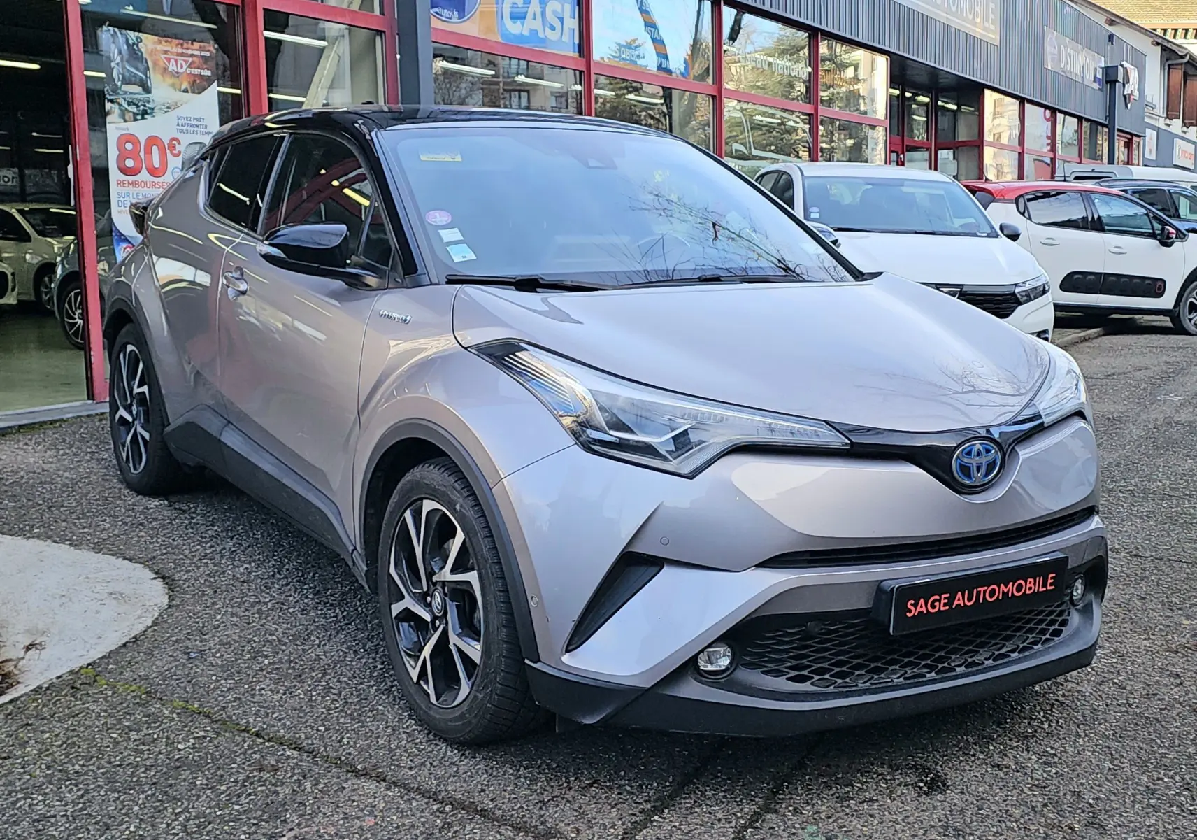 Vue 3/4 avant d'un Toyota C-HR hybride gris 2018 avec jantes noires et grises, stationné devant un garage.