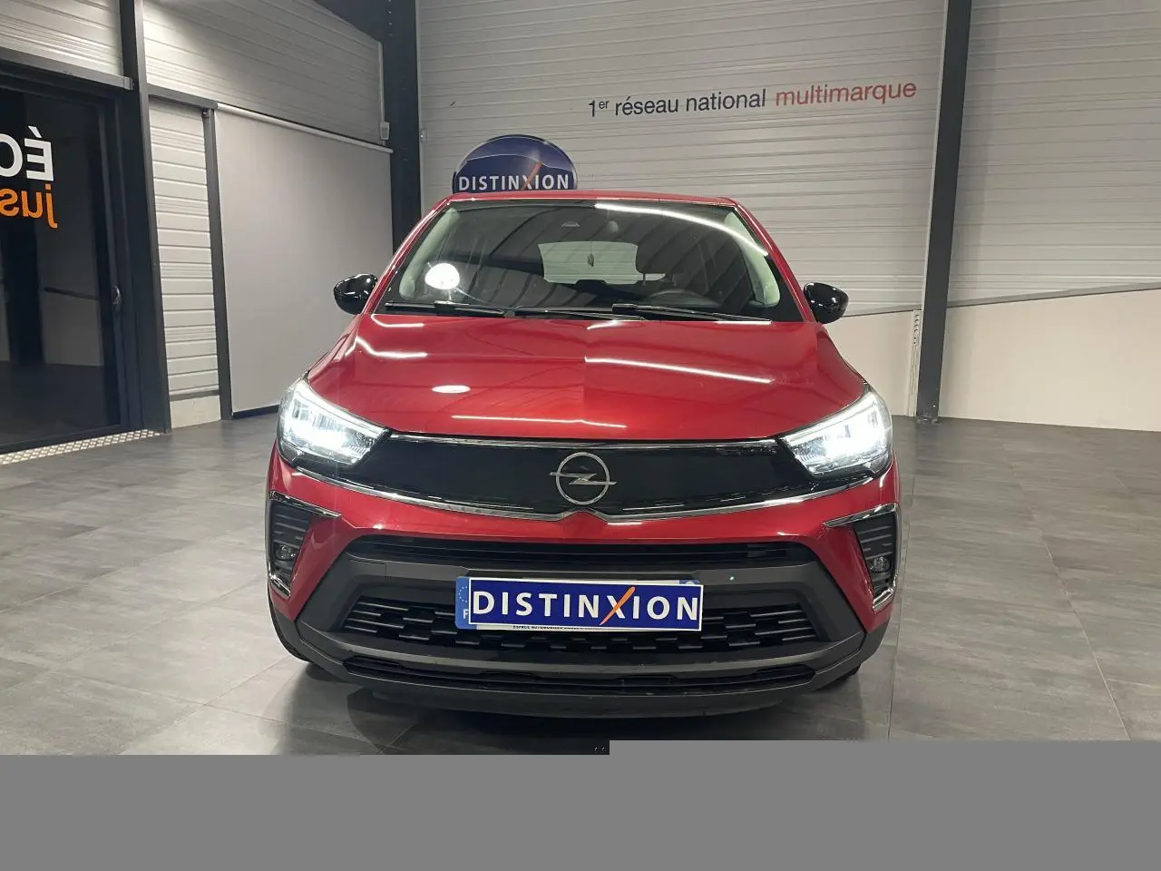 Vue de face de l’Opel Crossland rouge clair 2022 avec calandre noire et phares LED allumés en intérieur showroom.