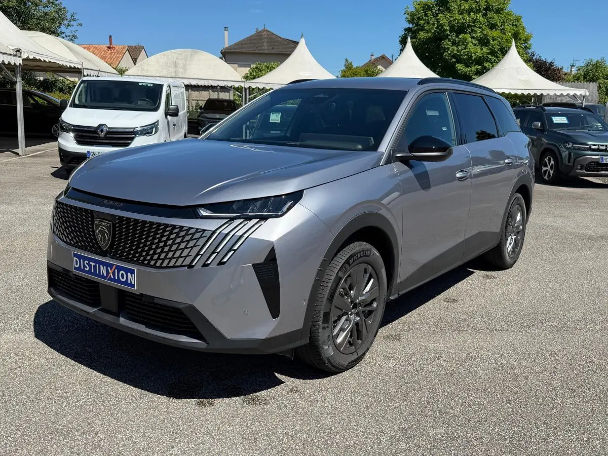 Peugeot 5008 gris clair en 3/4 avant droit, avec calandre distinctive et phares LED fins, sur parking extérieur.