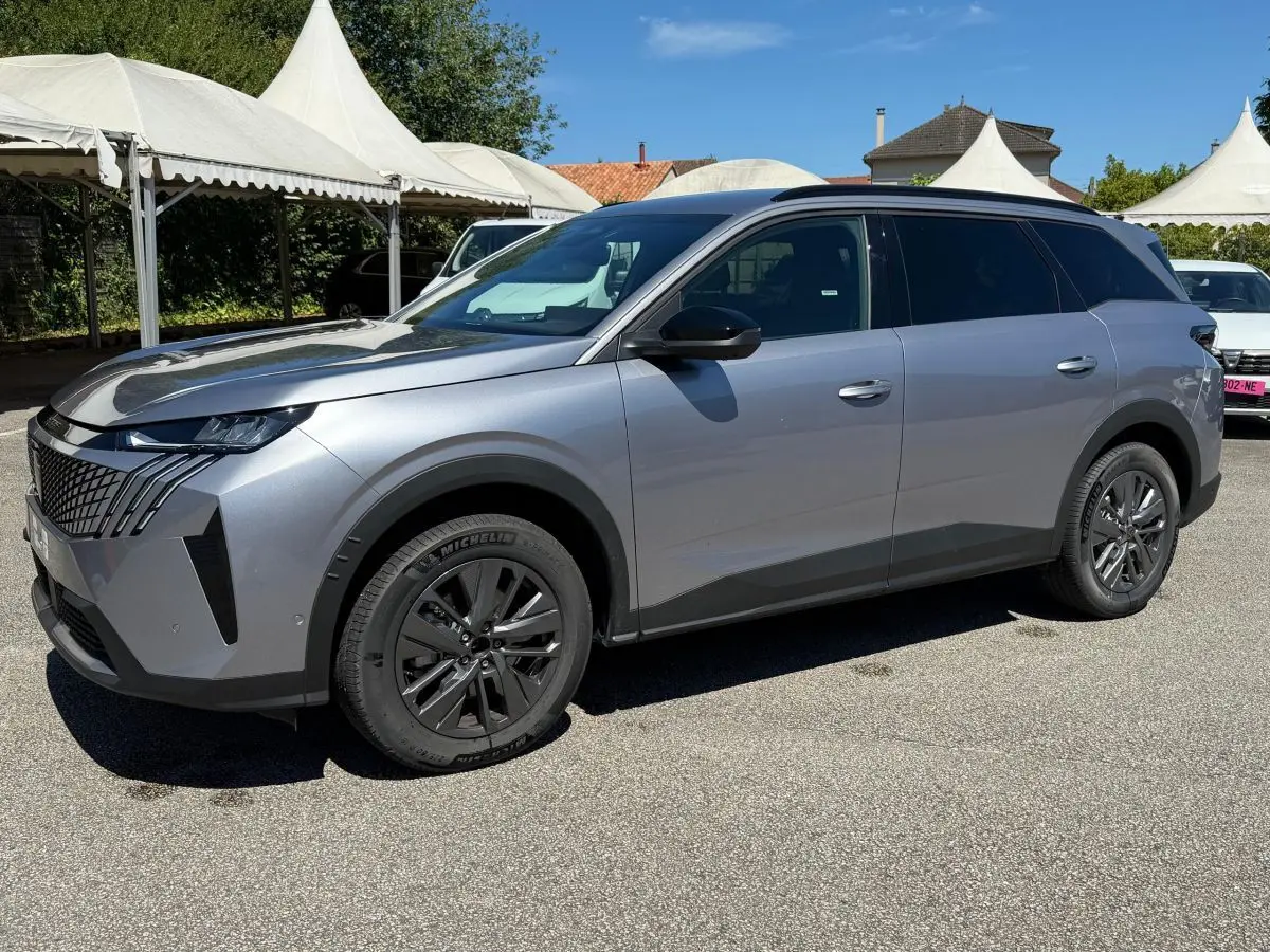 Peugeot 5008 gris clair vue 3/4 avant droit, avec calandre distinctive et jantes noires sur parking extérieur.