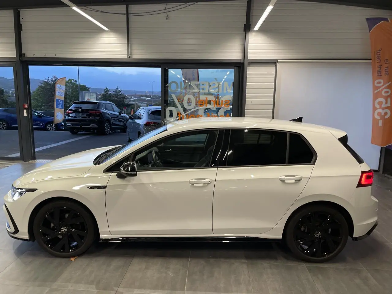 Vue de profil côté gauche d'une Volkswagen Golf blanche avec jantes noires en alliage et vitres surteintées en intérieur showroom.