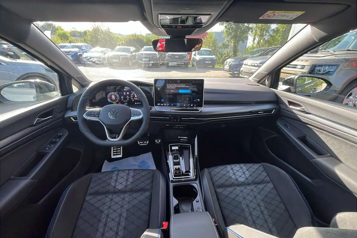 Vue intérieure avant de la Volkswagen Golf E-TSI 150 DSG R-Line avec tableau de bord numérique et sièges tissu noirs.