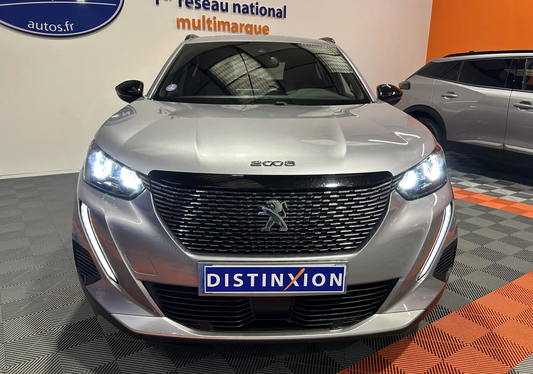 Vue avant d'un Peugeot 2008 gris Artense avec phares allumés et calandre noire brillante en intérieur showroom.
