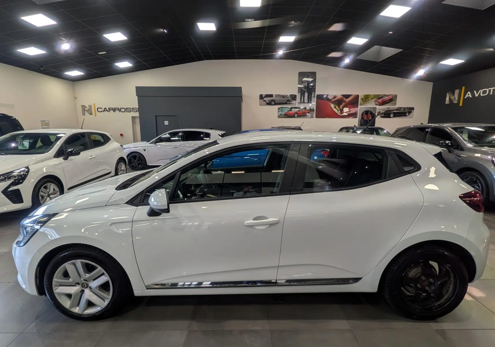 Profil côté gauche d'une Renault Clio V Business blanche Glacier, avec jantes différentes avant et arrière, en intérieur showroom.