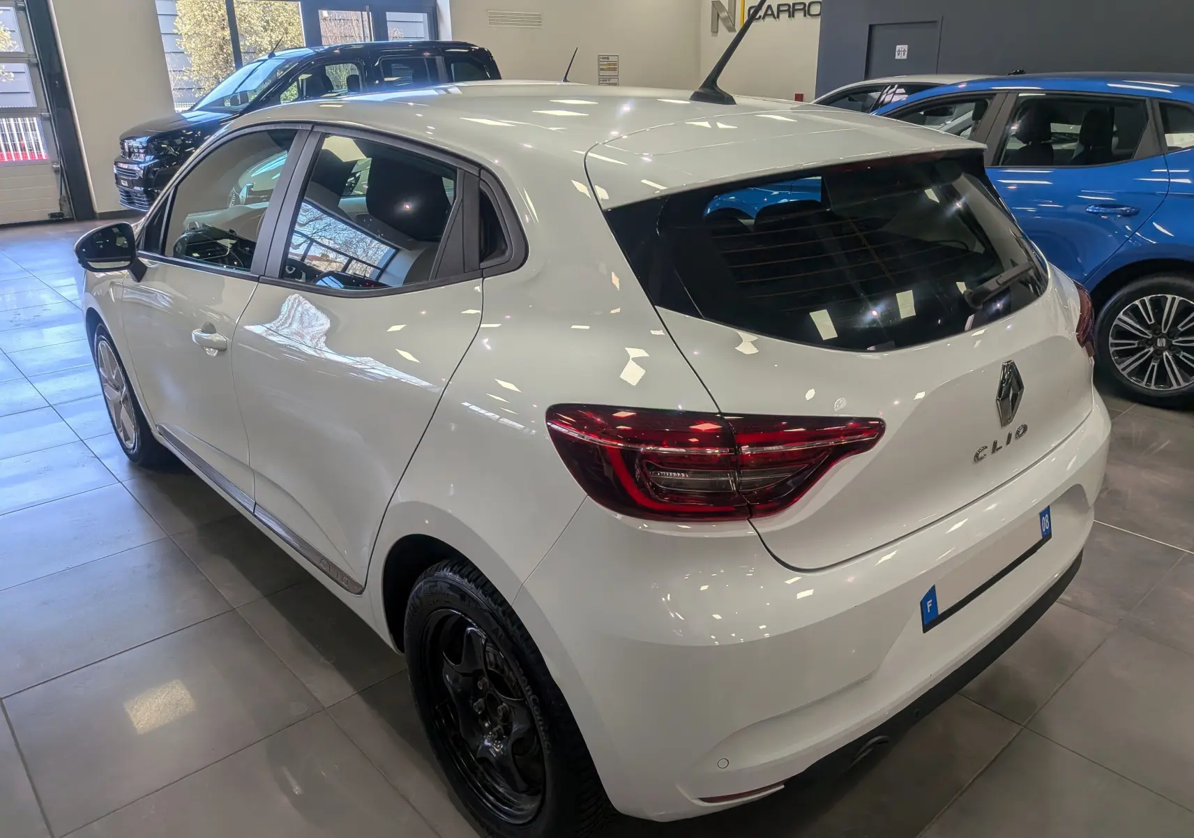 Vue 3/4 arrière droite d'une Renault Clio V Business blanc glacier en intérieur, avec feux arrière LED et jantes noires.