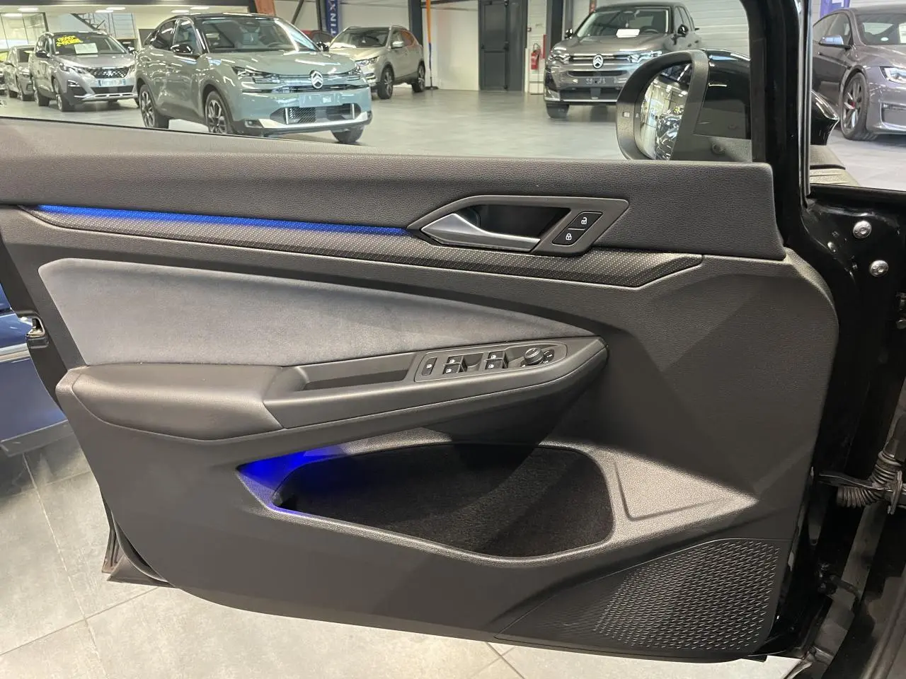 Vue intérieure côté gauche de la porte d’une Volkswagen Golf 2024 noire avec éclairage d’ambiance bleu et commandes électriques.