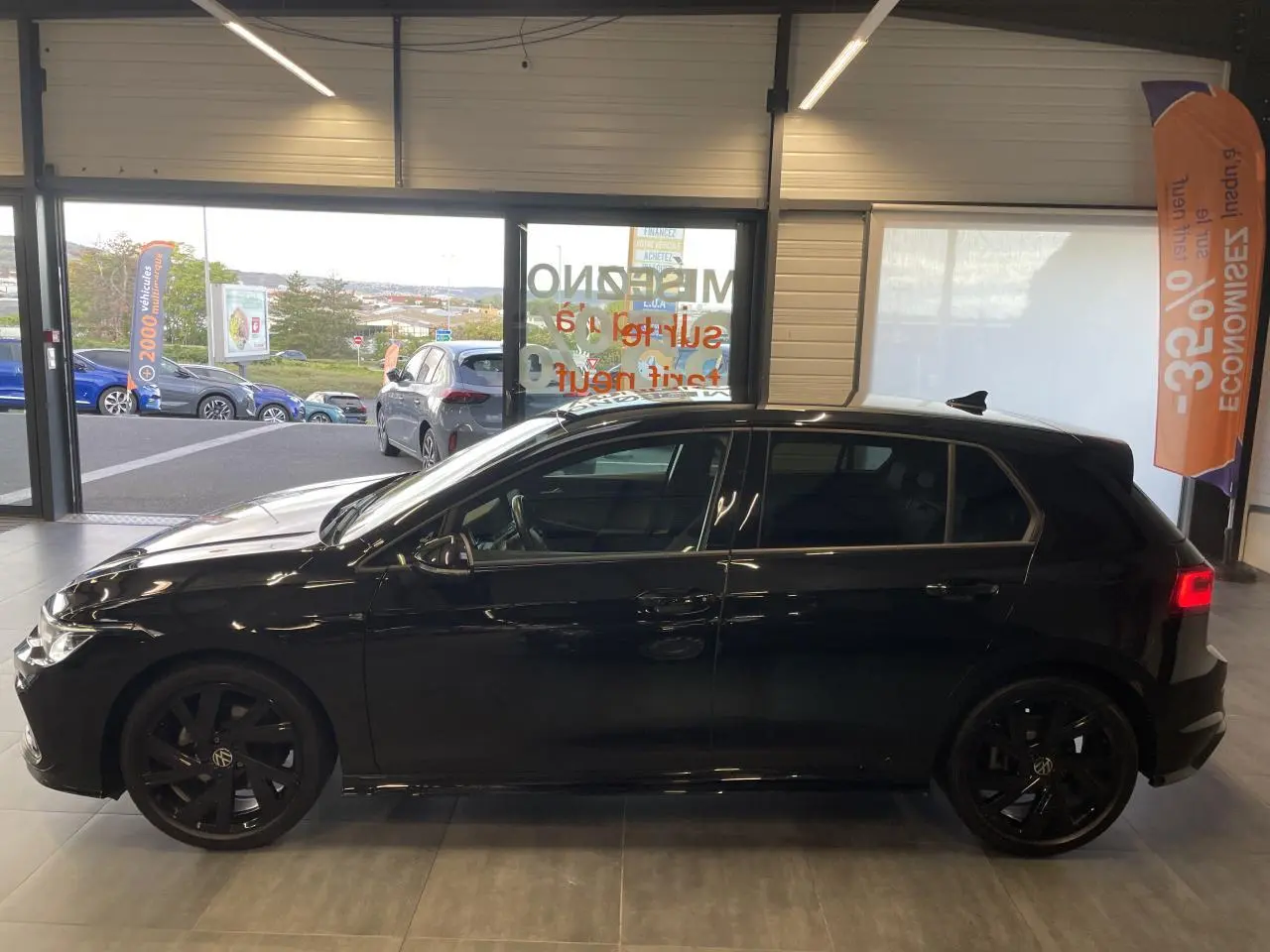Profil côté gauche d'une Volkswagen Golf 8 R-Line noire métallisée avec jantes Bergamo noires en intérieur showroom.