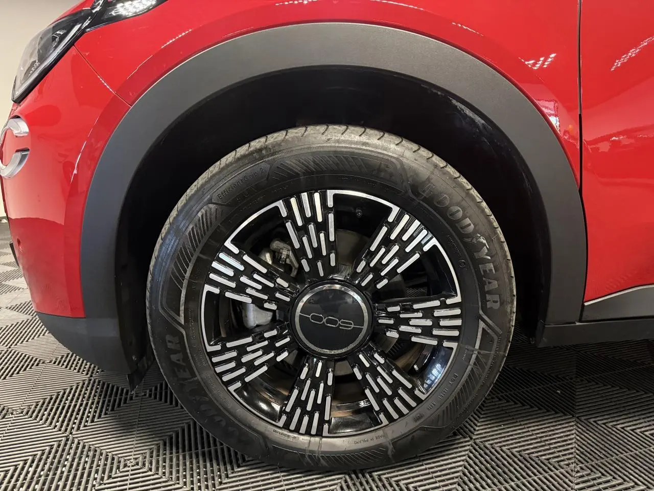 Gros plan sur la roue avant gauche d’une FIAT 600 rouge foncé avec jante noire et motifs argentés.