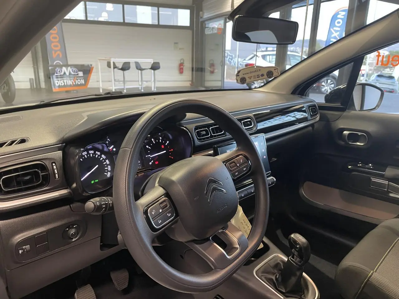 Vue intérieure côté conducteur du tableau de bord et volant noir d'une Citroën C3 beige clair 2024 avec boîte manuelle.