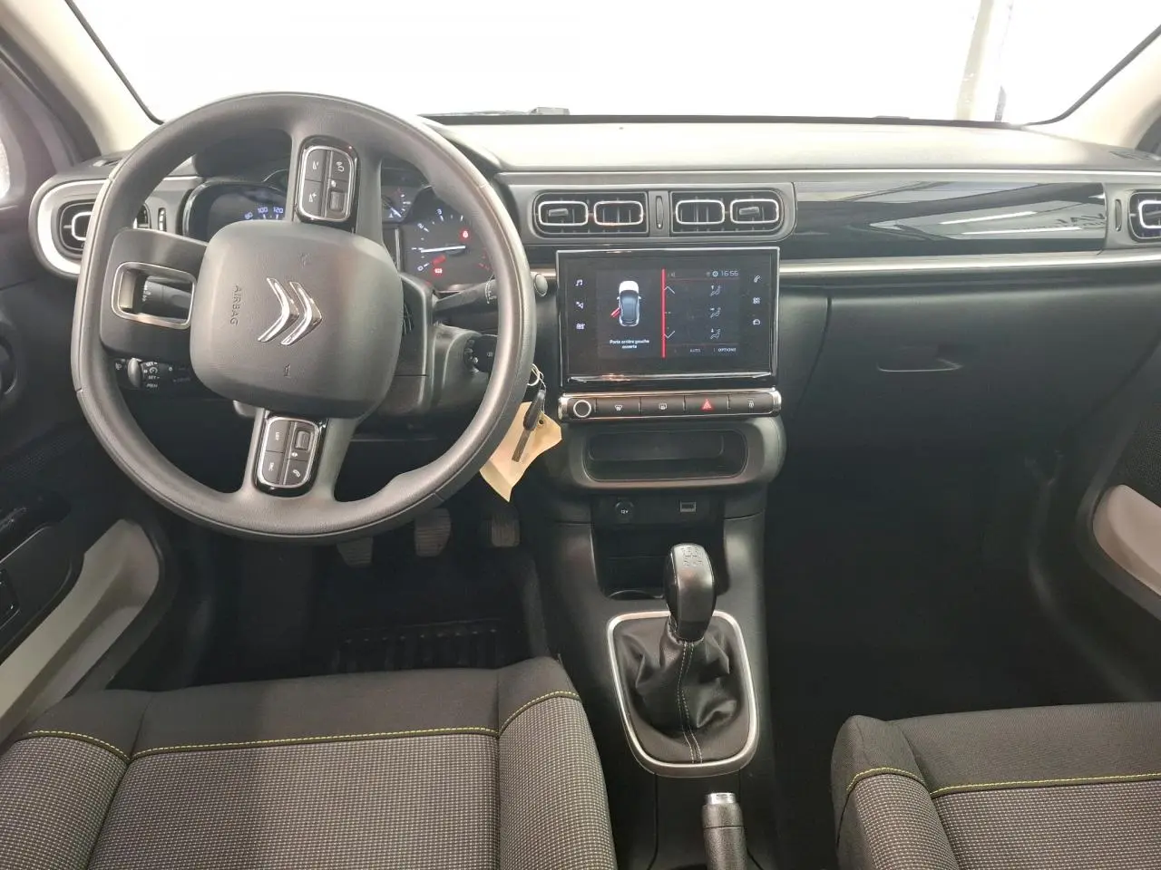 Intérieur de Citroën C3 2024 vu de face, tableau de bord noir brillant avec volant multifonctions et sellerie tissu gris à surpiqûres jaunes.