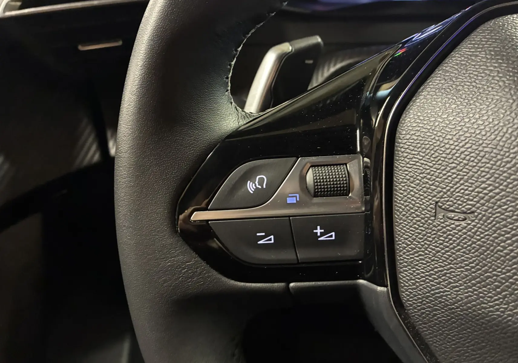 Gros plan sur les commandes gauche du volant noir du Peugeot 2008 1.2 Hybrid 136 bleu, avec molette et boutons lumineux.