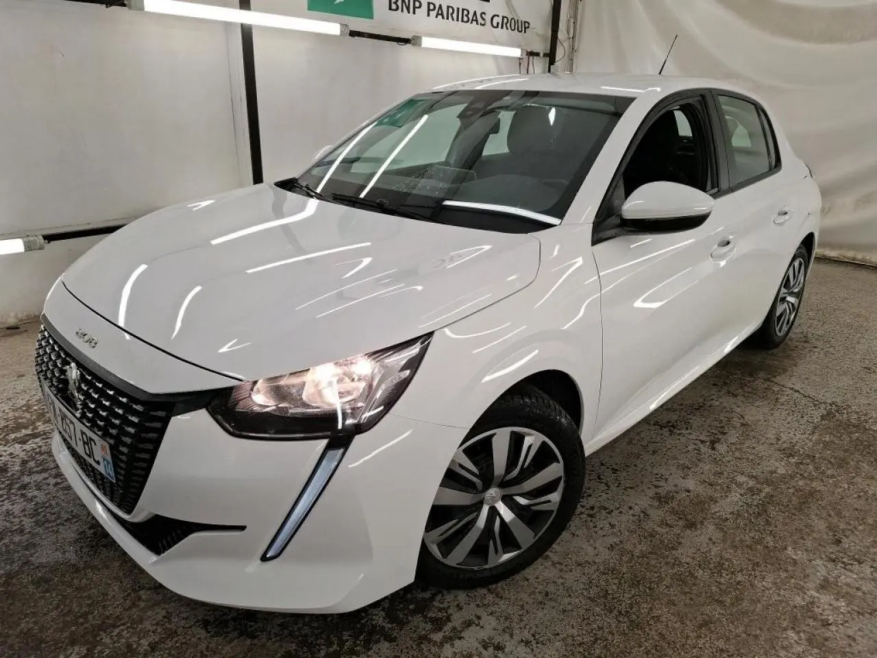 Peugeot 208 blanche vue en 3/4 avant droit, avec calandre noire et feux allumés dans un garage.