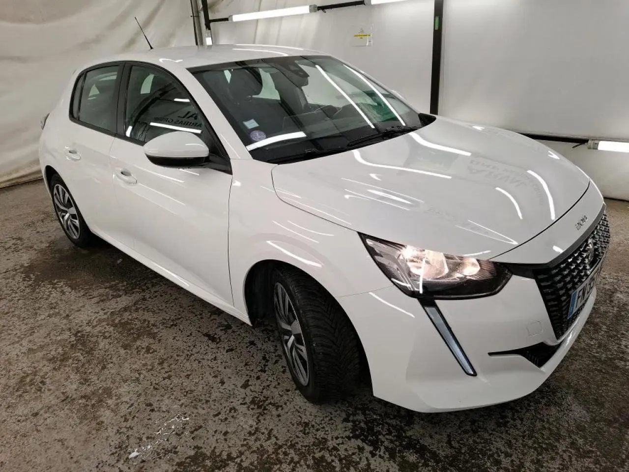 Peugeot 208 blanche vue en 3/4 avant droit, avec calandre noire et feux allumés dans un garage.