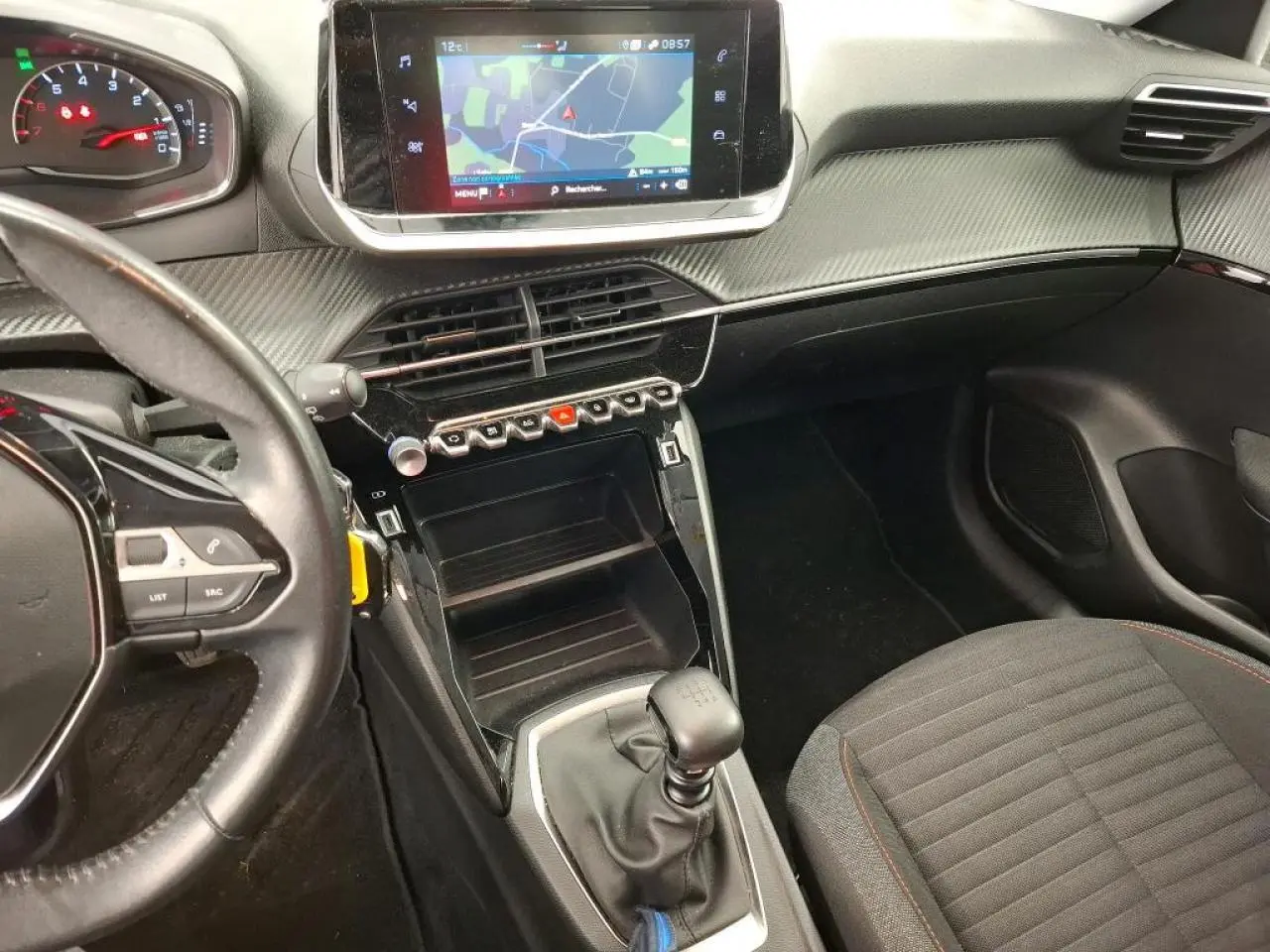 Vue intérieure côté conducteur de la Peugeot 208 blanche, avec tableau de bord carbone et écran tactile central.