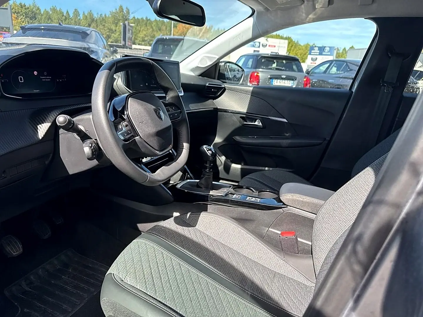 Intérieur avant droit de la Peugeot 208 1.2 100 Allure 2025, volant compact et sièges tissu gris, avec levier de vitesse manuel.