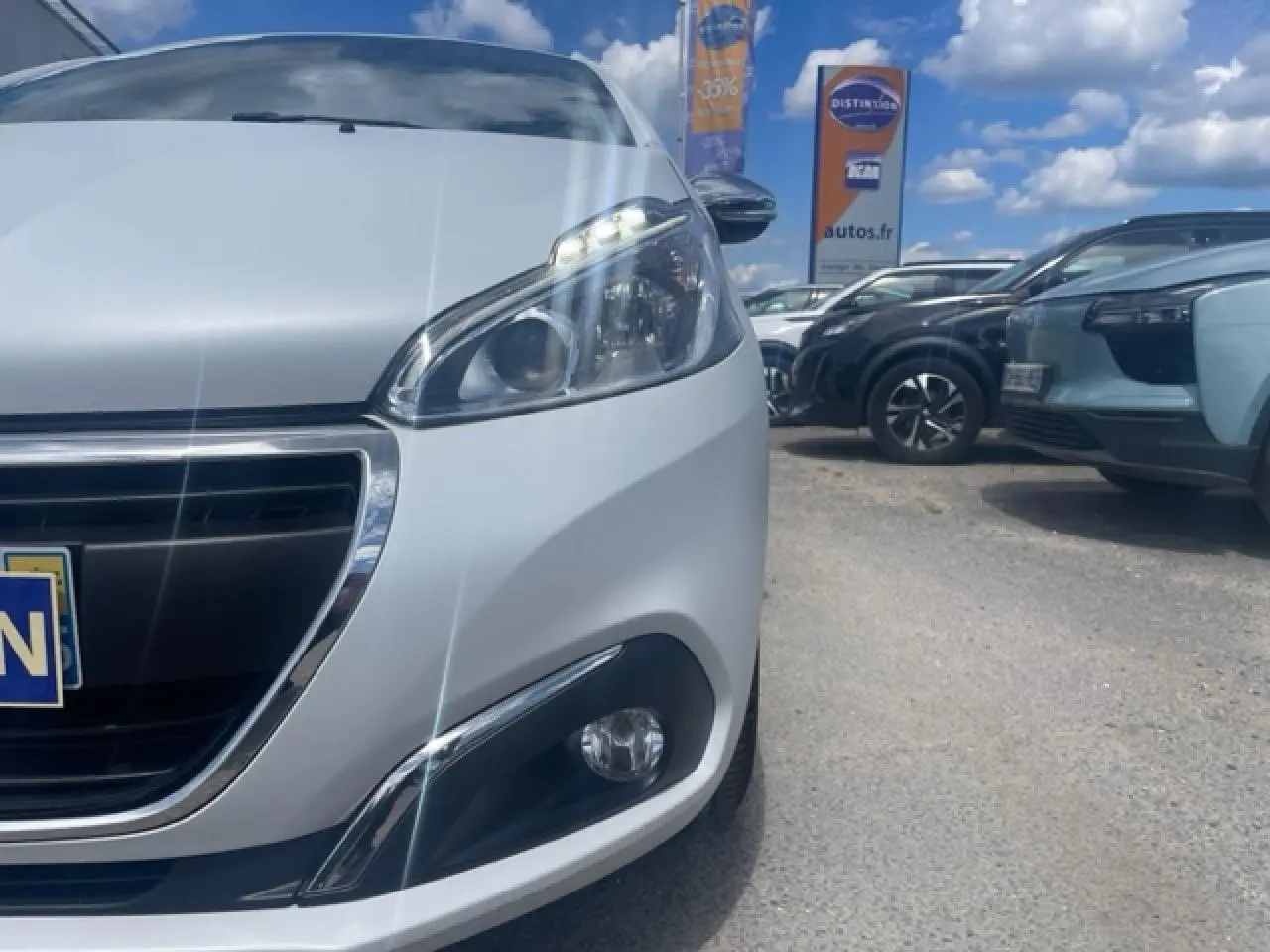 Gros plan sur l'avant droit d'une Peugeot 208 blanc perle avec phares et calandre chromée sous un ciel bleu.