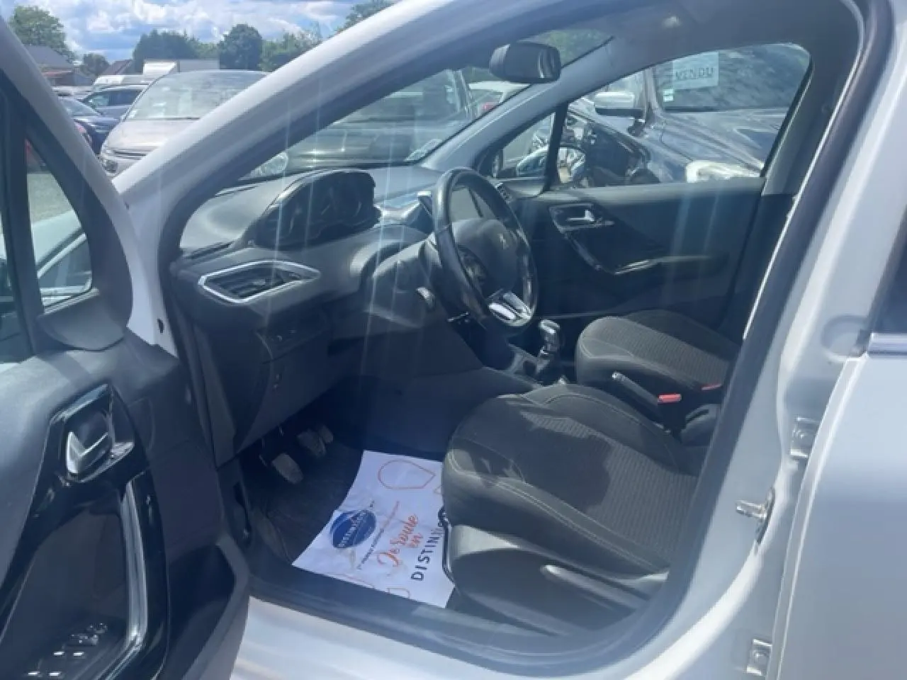 Vue intérieure côté conducteur d'une Peugeot 208 blanc perle, montrant les sièges noirs et le volant multifonction.