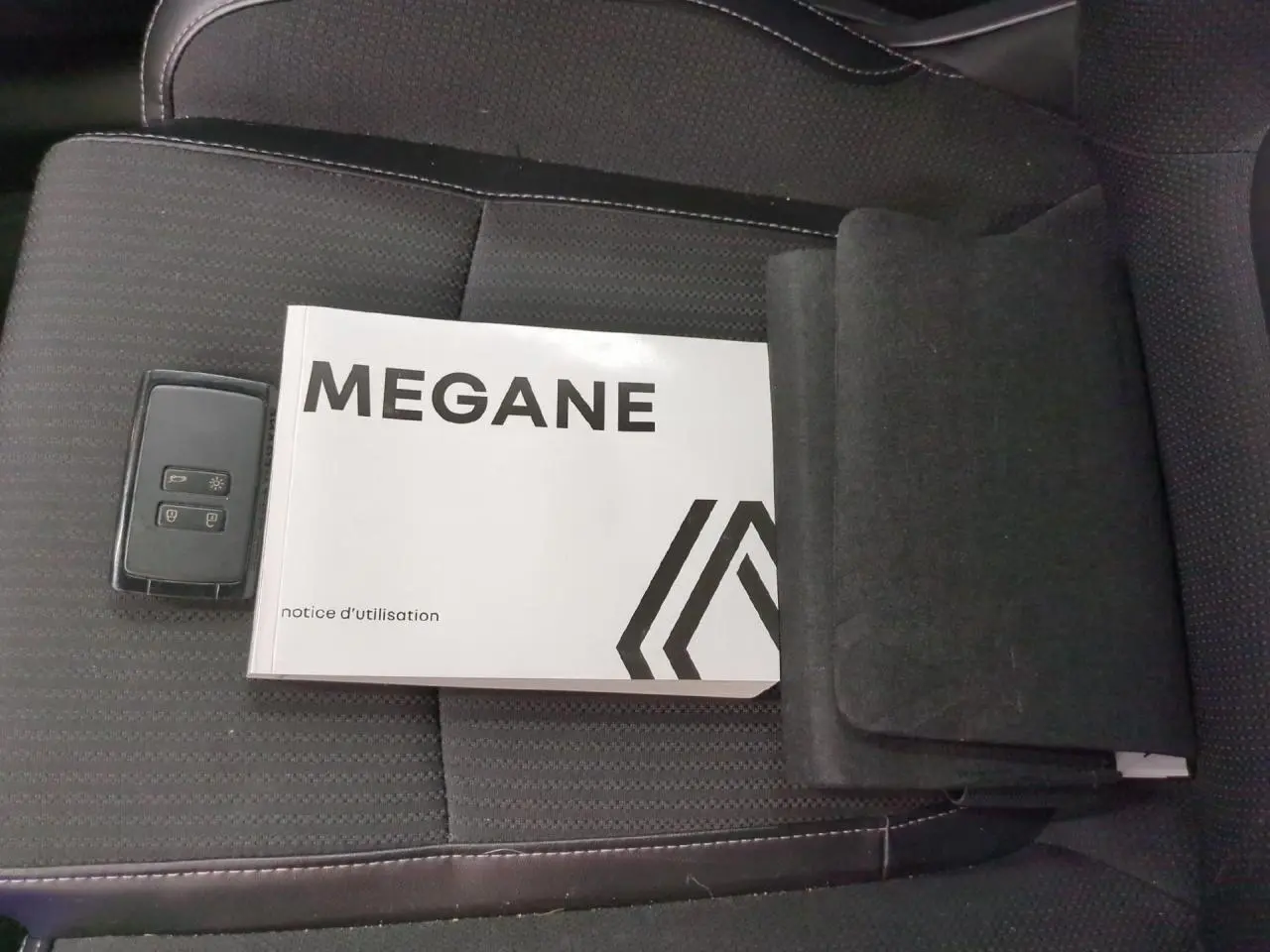 Notice d'utilisation Mégane et clé Renault posées sur un siège avant en tissu noir avec surpiqûres grises.