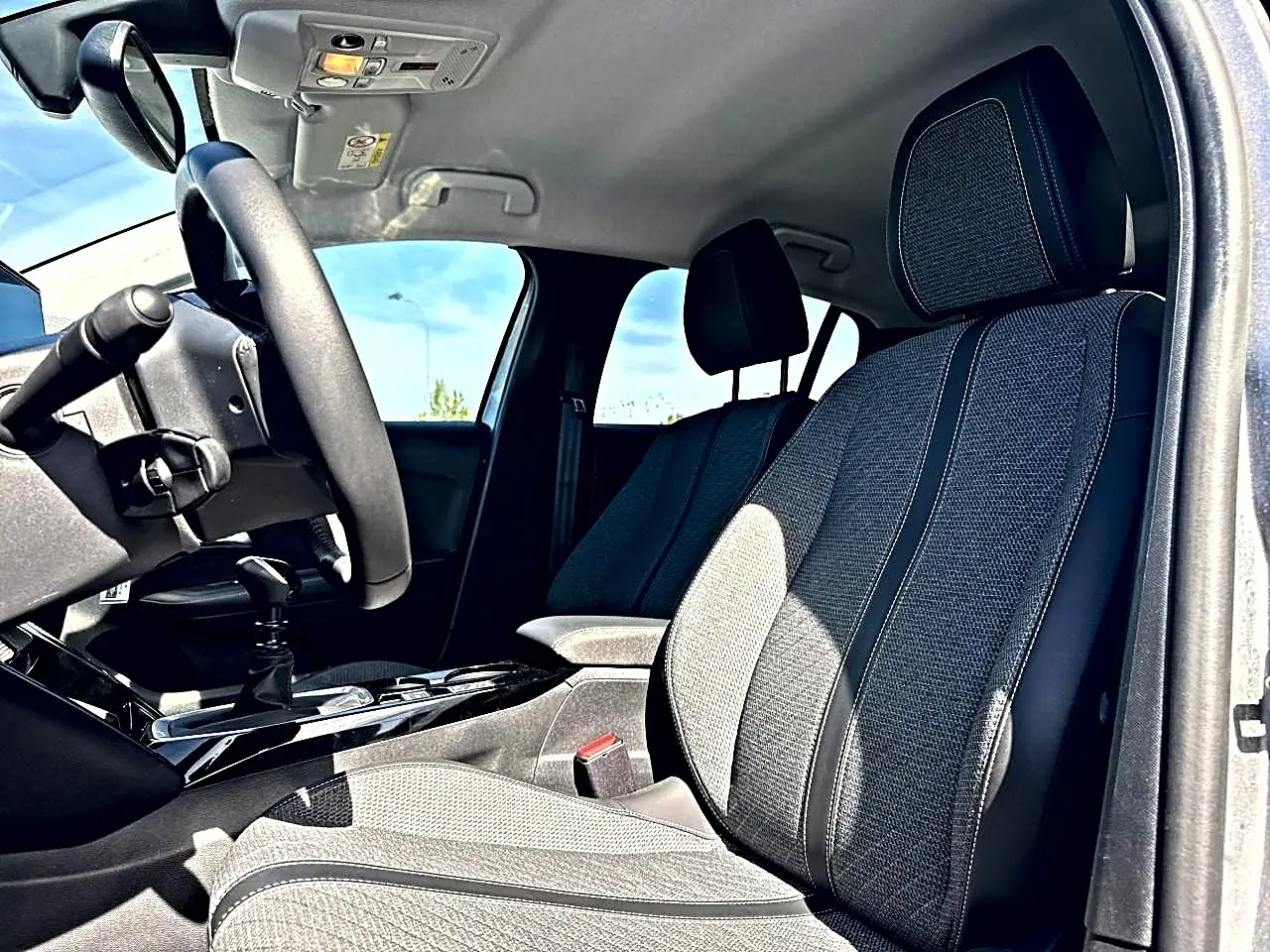 Vue intérieure côté conducteur de la Peugeot 208 1.2 100 allure 2025, sièges tissu gris et volant noir.