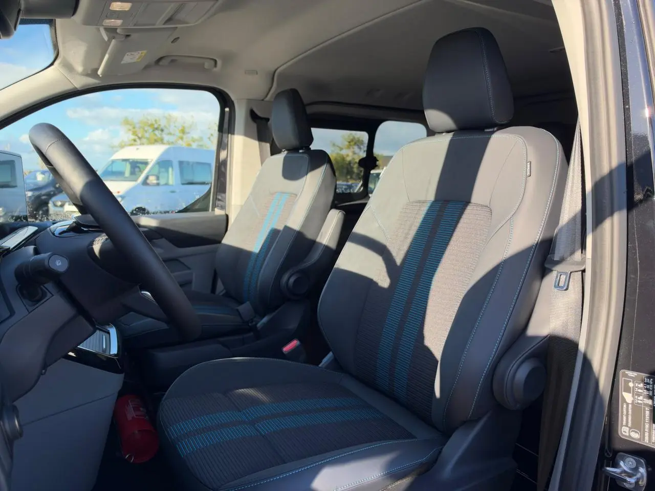 Vue intérieure côté conducteur du Ford Transit Fourgon 2026, sièges sport gris avec bandes bleues et volant noir.