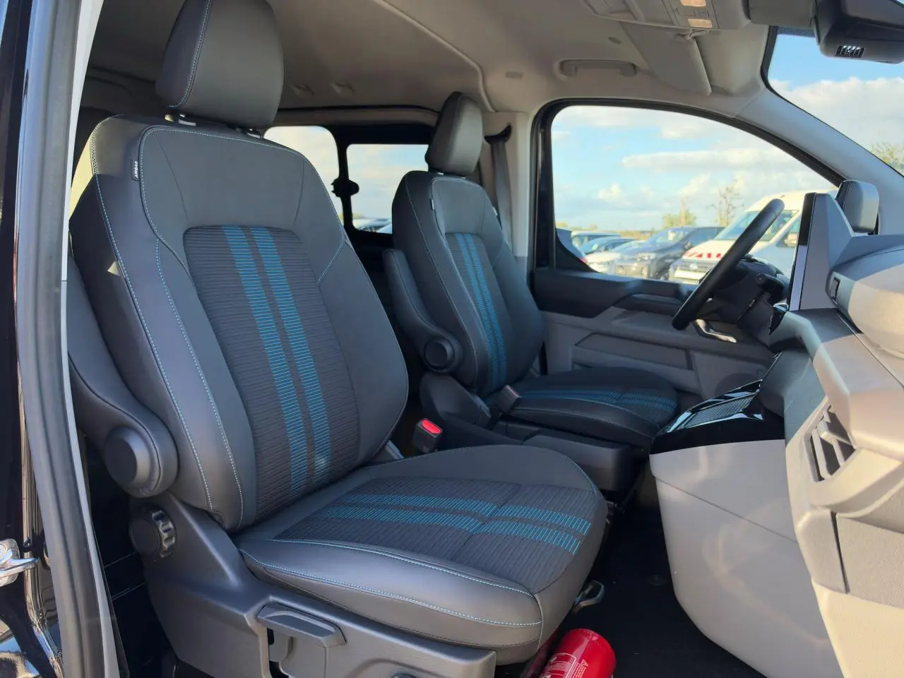 Vue intérieure côté conducteur du Ford Transit Fourgon 2026, sièges sport noirs avec bandes bleues et tableau de bord gris.