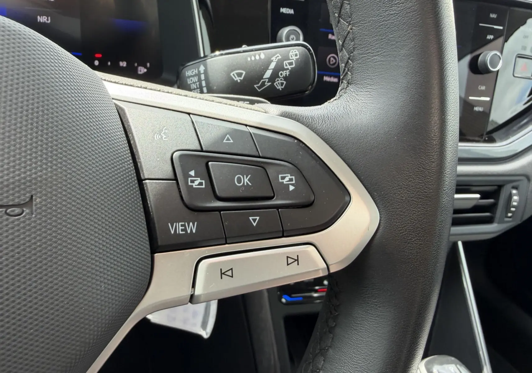 Gros plan sur les commandes multifonctions en cuir noir du volant de la Volkswagen Polo 2025, avec écran tactile en arrière-plan.