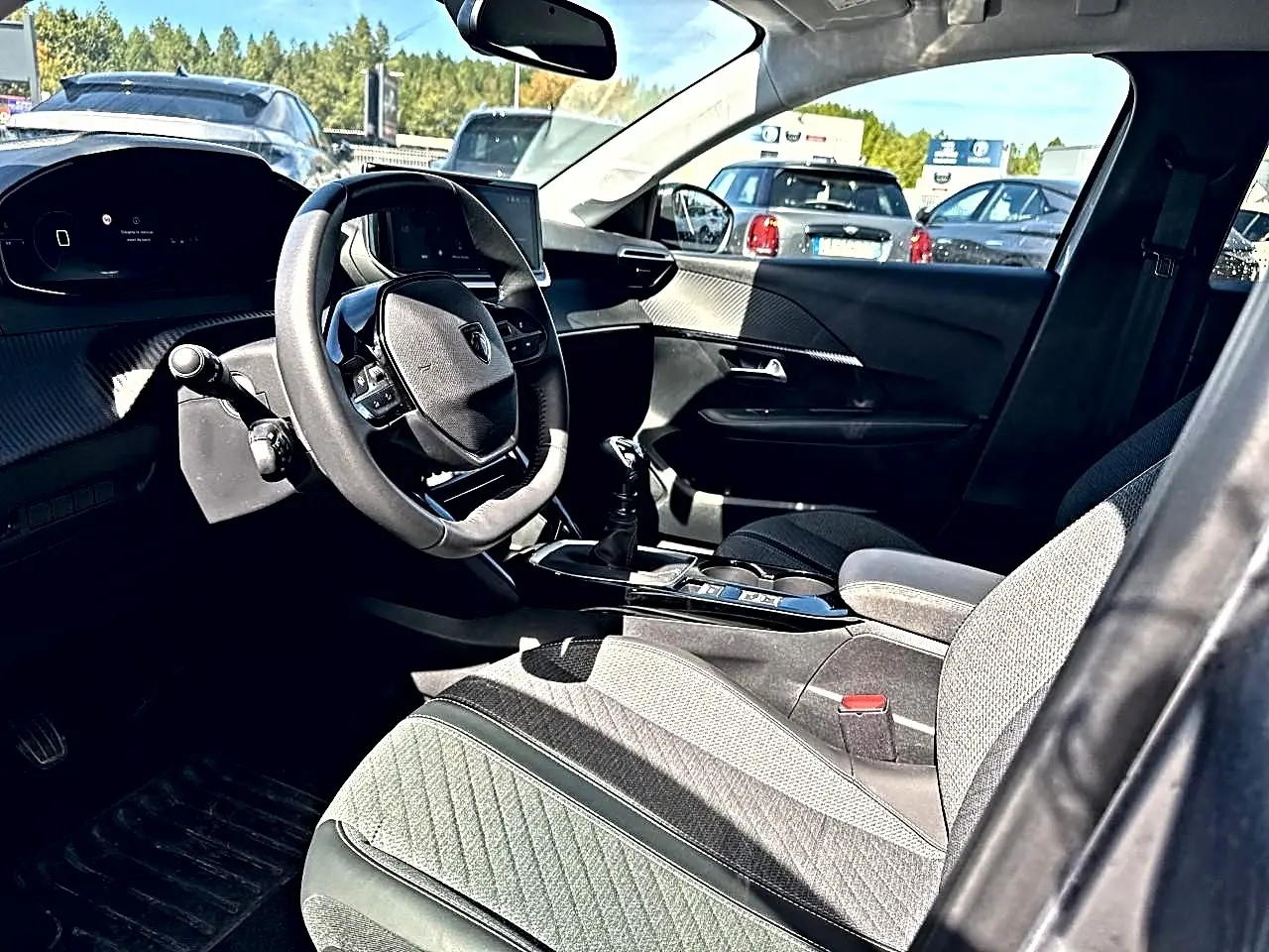 Vue intérieure côté conducteur de la Peugeot 208 1.2 100 Allure 2025 avec volant compact et sellerie tissu gris.