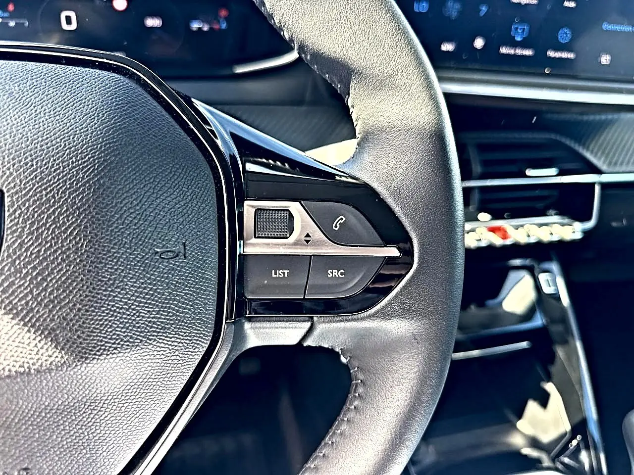 Gros plan sur le volant noir de la Peugeot 208 1.2 100 Allure avec commandes audio intégrées, tableau de bord numérique en arrière-plan.