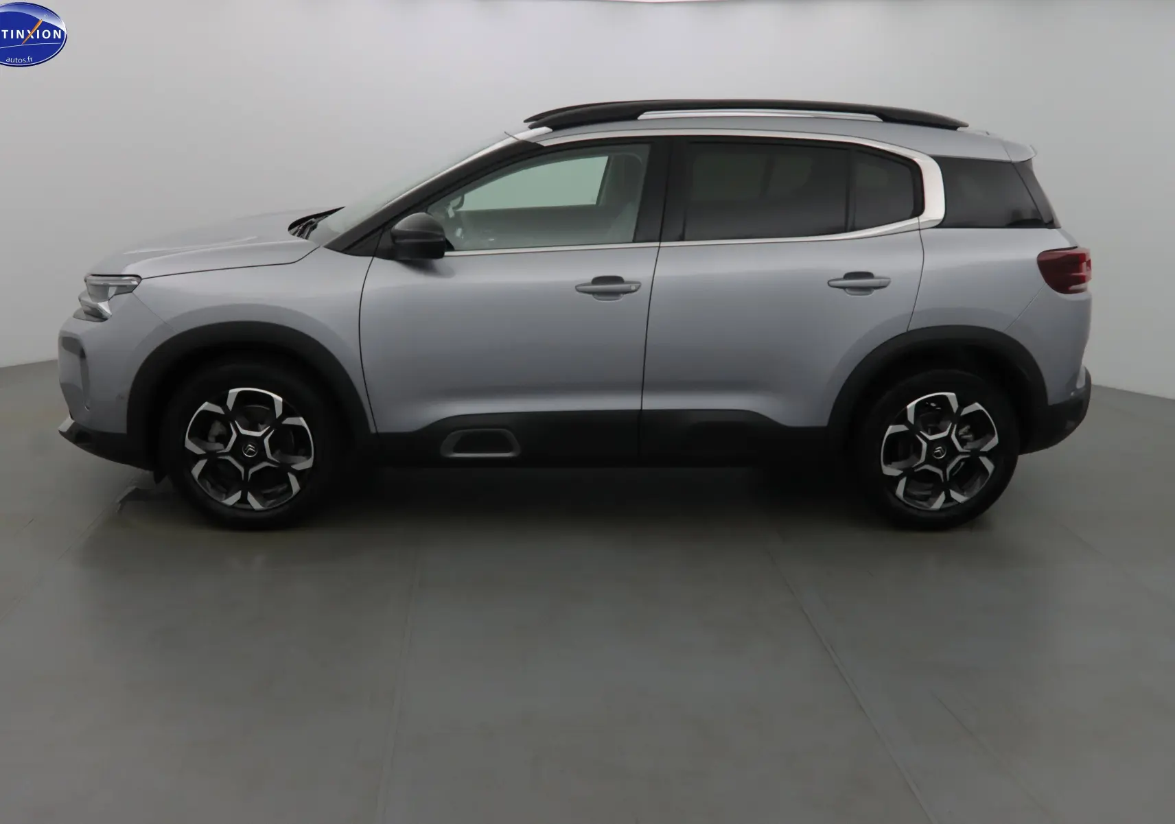 Profil latéral gauche d'un Citroën C5 Aircross gris Artense métal avec barres de toit et protections noires sur les passages de roues.